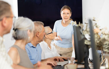 Adult female teacher showing group of elderly people how to work with computer at computer course