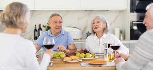 Two mature married couple sitting at table, enjoying food and wine, chatting, talking, laughing. In...