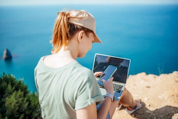 A woman is sitting on a rock by the ocean, using her laptop and cell phone