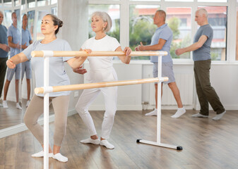 Elderly active women perform various ballet positions at the barre, men in the background perform...