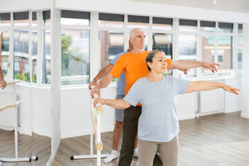 Mature woman in sportswear participating 2nd position of ballet stance at barre with group in bright choreography studio