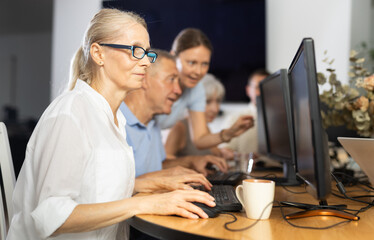 Focused senior woman in glasses attending group computer class, highlighting interest and...