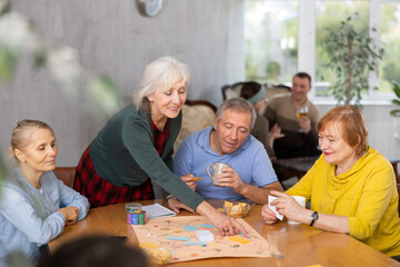 Smiling elderly woman with group of friends sitting together around desk, playing entertaining...