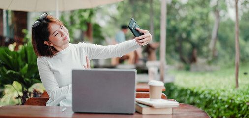 A woman is taking a selfie with her cell phone while sitting at a table with a laptop and books