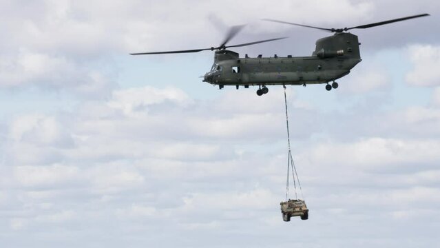 An RAF Chinook helicopter hovering in the open sky while lifting an armoured personnel carrier