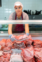 Smiling young girl, saleswoman of butcher shop, arranging meat products in glass display case, laying out slabs of fresh raw pork ribs