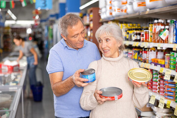Attentive man and woman purchasers choosing tinned fish out of large stock in a big supermarket