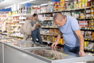 Positive mature man shopping in supermarket, choosing frozen convenience food