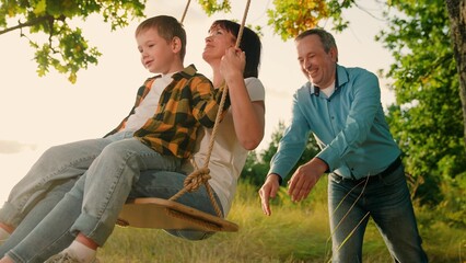 Son child, dad, mom play together on wooden swing, dreams of flying. Children swing. Father, mother...