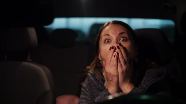 Curious woman looks with intense attention widening eyes sitting in car. Lady driver expresses fear screaming and closes mouth with hands. Girl watching a movie in a drive-in cinema