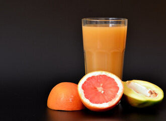 Fruit juice in a tall glass on a black background, next to pieces of ripe grapefruit and mango.