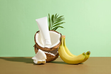 Two halves of dried coconut are stacked on top of each other with a white cosmetic tube without brand above beside a bunch of yellow banana on brown table top over green texture
