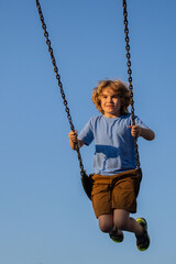 Kid swinging on chain swing on city kids playground. Swing ride. Cute child having fun on a swing on summer sky background. Blonde little boy swings at kid playground. Child swinging high.