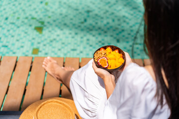 Young woman traveller relaxing and eating refresh bowl with fruits at tropical resort pool on summer vacation