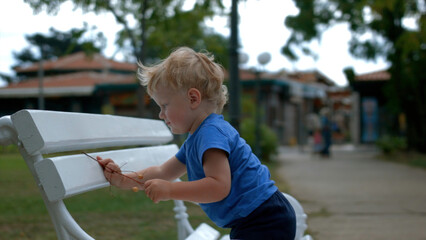 Cute baby boy in climbing on the bench in a city park. Creative. Kid crawls on white bench on a...