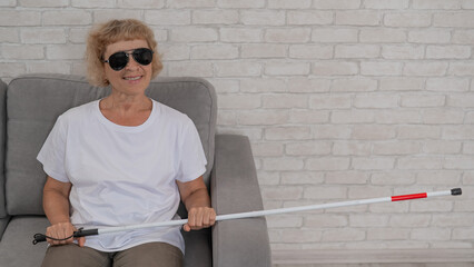 An elderly blind woman wearing sunglasses and with a tactile cane sits on the sofa. 