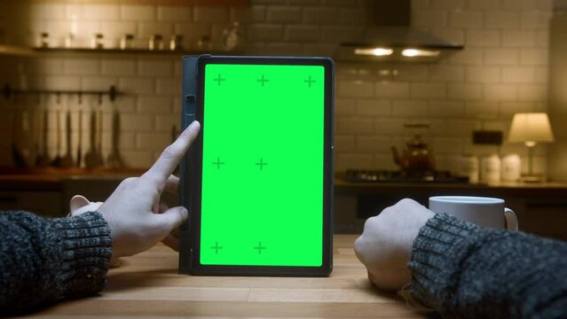 Point of view shot: Close-up of man using green mock-up screen digital tablet computer on the table. Man scrolls and taps on tablet computer in modern kitchen background.