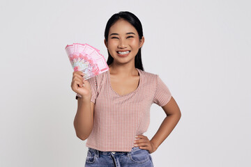 Smiling happy young Asian woman wearing a casual t-shirt holds in hand fan of cash money in rupiah...