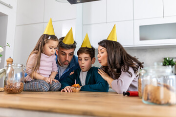 In the kitchen, a father blows out birthday candles on a cake, surrounded by his happy family....