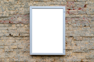 Blank white mockup background texture of a poster with a metal frame mounted on a rough and grungy brick wall. An empty vertical advertisement or notice board on an old building.