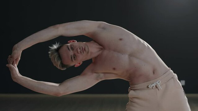 Slowmo medium shot of shirtless slim Caucasian young man stretching his back standing on parquet floor against black wall
