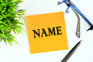 A word name on a yellow sticker on a light background next to glasses, a pen and a green plant