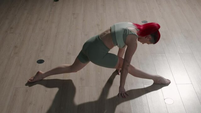From above slowmo of flexible red haired Caucasian young woman in tight activewear doing splits on parquet floor