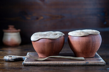 Two clay pots with stewed vegetables on a wooden table, closeup. Stewing food in earthenware is...