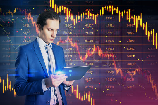 Attractive young european businessman using tablet with downward red forex candlestick hologram on blurry office interior background. Financial crisis, stock and recession concept. Double exposure.