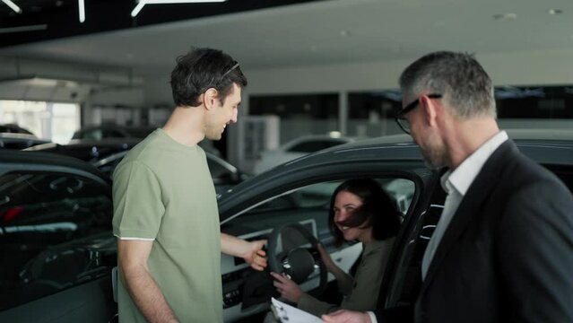 A happy brunette guy in a Green T-shirt talks to a manager a man with gray hair in a business suit about a car in a car showroom. A young couple a guy and a brunette girl in green T-shirts are talking