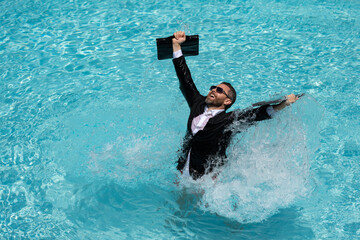 Business man in suit with laptop excited jumping in swimming pool. Summer travel tourism and business concept. Crazy office employee using laptop in pool on summer day.