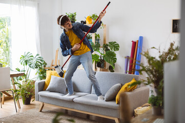 Happy man wearing wireless headphones and playing guitar on broom at home