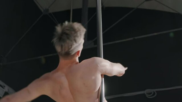 Low angle slowmo shot of shirtless Caucasian guy with blond hair spinning gracefully on pole indoors