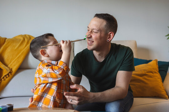 Boy painting happy fathers face with paint brush on sofa at home