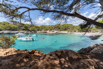 Image framed by lush vegetation and rocky terrain with serene turquoise waters of Cala Turqueta in...