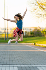 Happy young woman in blue tank top and red plaid shirt joyously leaping with headphones, arms spread wide, in a sunny urban park during sunset. 