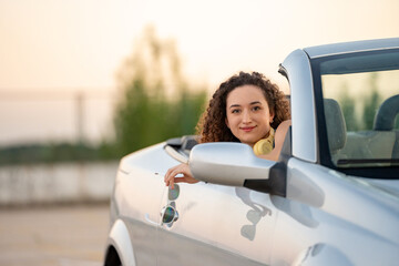 Relaxed young woman with curly hair and headphones enjoys a tranquil sunset from a convertible car. 