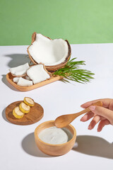 A hand holds a wooden spoon preparing to stir white cream in a wooden bowl placed on a white table top, behind is a wooden tray decorated with a few pieces of banana and a tray of coconut