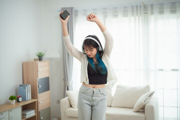 young woman with vibrant blue-tipped hair dances energetically in a modern living room. She wears...