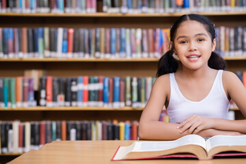 girl in the library. little Asian teenage girl sitting at the table with an open book in a white T-shirt against the background of a wall with books, reading concept