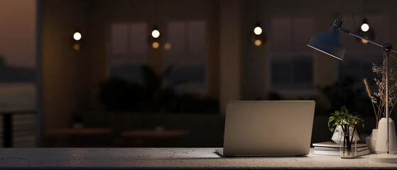 A table features a laptop computer and a dim light from a lamp in a dark coffee shop at night.