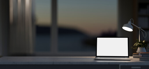 A white-screen laptop computer mockup on a table near the window in a modern dark room.