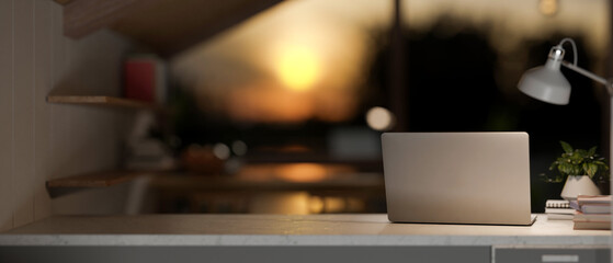 A laptop computer on a table in a room with dim lights from a lamp and sunset through the window.