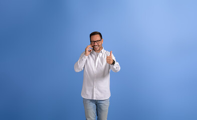 Portrait of confident businessman talking over phone and showing thumbs up sign on blue background