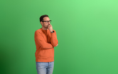 Young serious businessman with hand on chin thinking business ideas on isolated green background