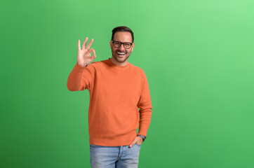 Portrait of confident salesman with hand in pocket showing OK sign while posing on green background