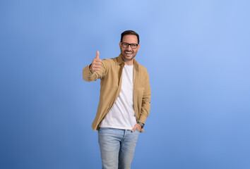 Young businessman with hand in pocket smiling and gesturing thumbs up sign against blue background