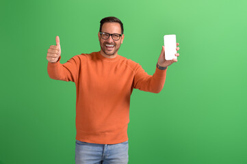 Cheerful young salesman showing thumbs up sign and smart phone's screen on isolated green background