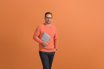 Portrait of serious businessman with hand in pocket holding wireless computer on orange background