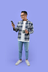 Full-length Portrait of a young cheerful excited Asian man standing isolated over a purple background, using a mobile phone while holding a cup